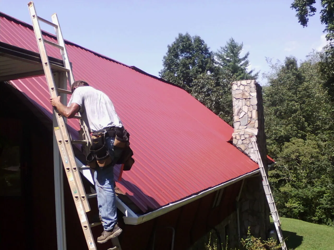 Roofer on a ladder beside a red metal roof and stone chimney — Soffit & Fascia Repair by Roofing Brothers in Hersey